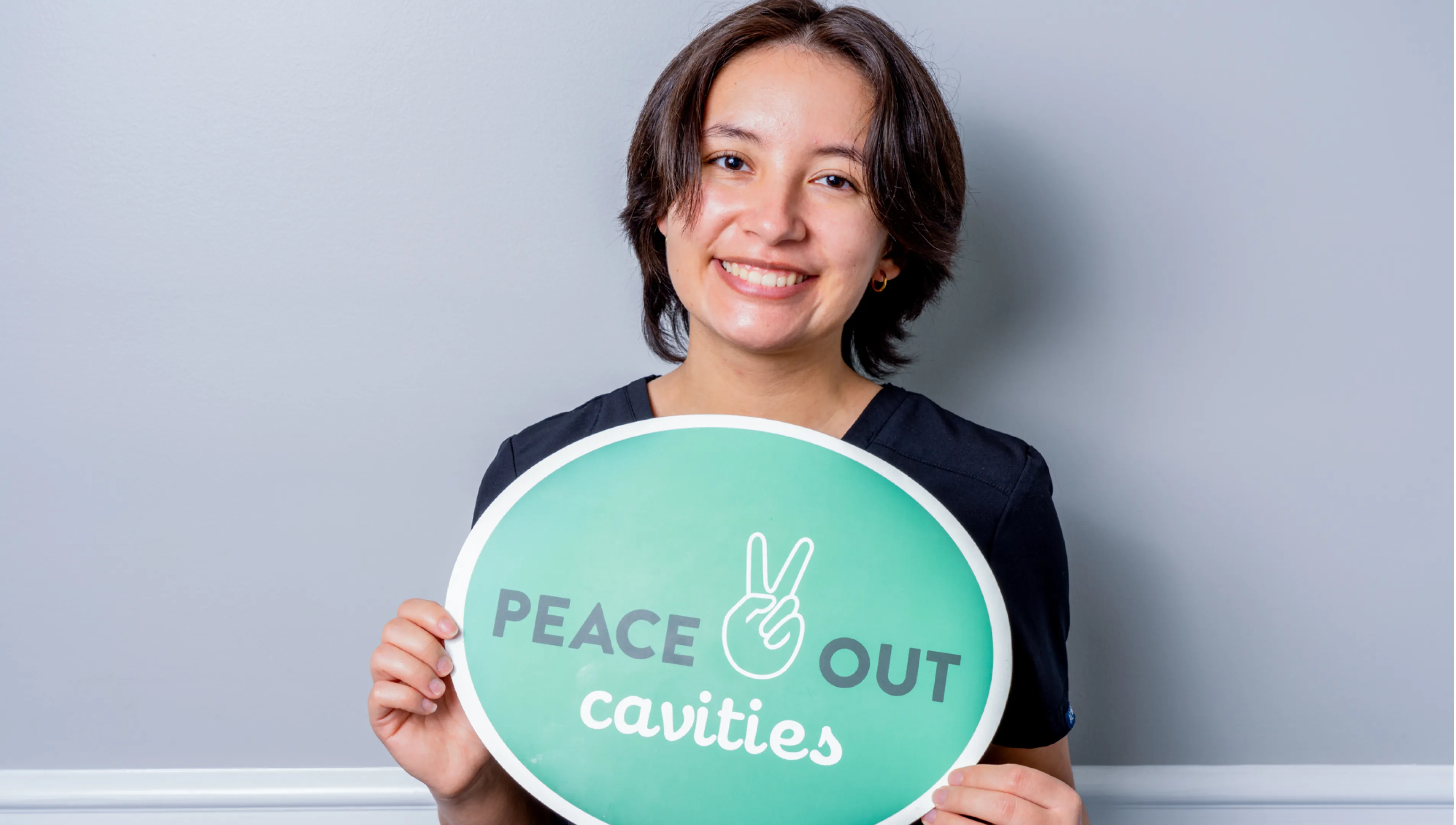 Smiling woman holding a green oval sign that says 'PEACE OUT cavities' with a hand peace symbol.